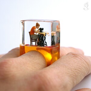 Op de afbeelding: Een unieke ring met een miniatuursc&egrave;ne van een persoon die naait aan een vintage naaimachine, ingekapseld in een helder, rechthoekig harsblok. De ring heeft een amberkleurige basis en wordt om een vinger gedragen.