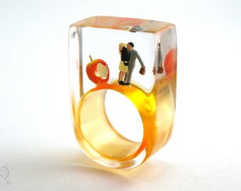 Warm-hearted – paradisiacal lovers ring with kissing mini figures next to an apple on an orange ring made of resin for lovers