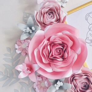 May include: A paper flower arrangement with large pink roses, smaller pink flowers, and gray leaves. The flowers are layered and create a 3D effect.