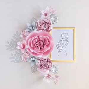 May include: A framed line drawing of a mother holding a child, surrounded by pink and gray paper flowers. The flowers are arranged in a cluster and include roses, butterflies, and leaves.