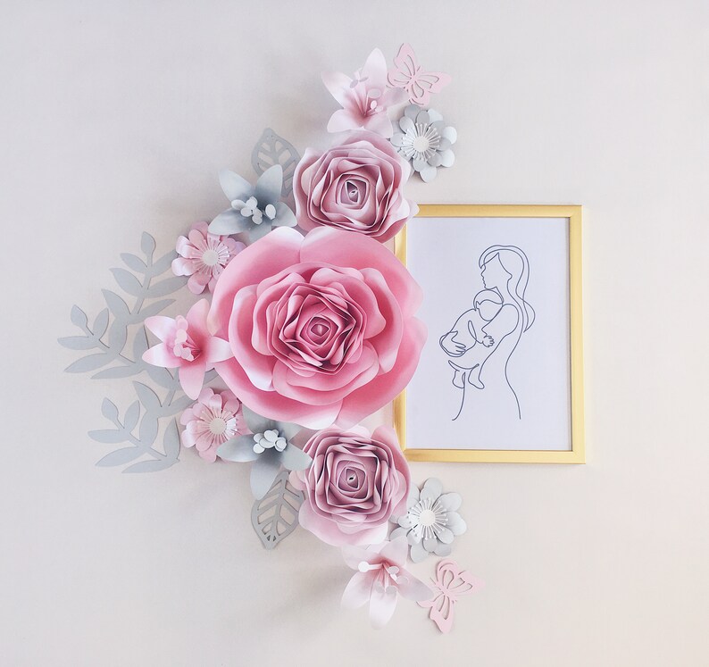 May include: A framed line drawing of a mother holding a child, surrounded by pink and gray paper flowers. The flowers are arranged in a cluster and include roses, butterflies, and leaves.
