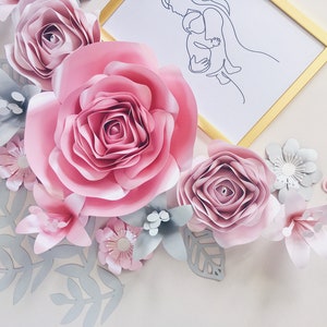 May include: A paper flower arrangement with a pink, white, and gray color scheme. The arrangement features a large pink rose, smaller pink roses, white flowers, and gray leaves. A framed line drawing of a mother and child is in the background.