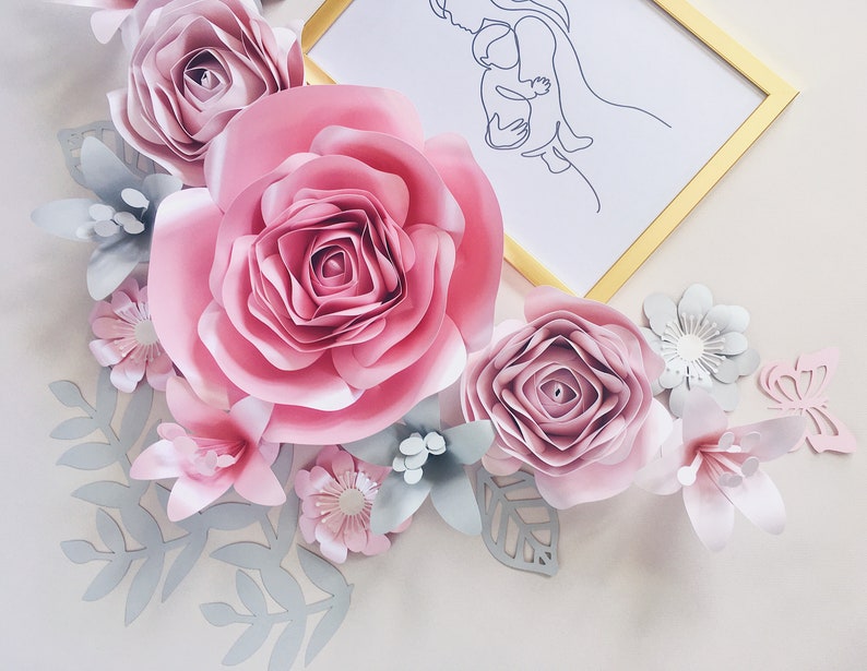 May include: A paper flower arrangement with a pink, white, and gray color scheme. The arrangement features a large pink rose, smaller pink roses, white flowers, and gray leaves. A framed line drawing of a mother and child is in the background.