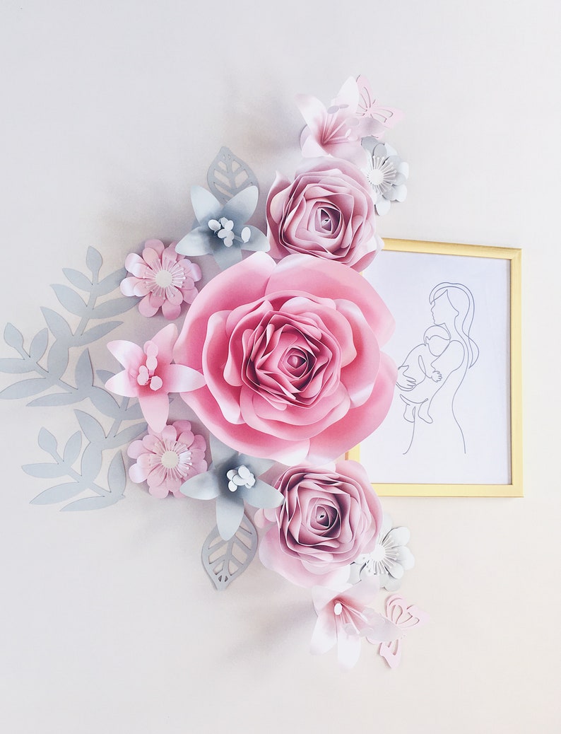 May include: A wall decor featuring a paper flower arrangement in shades of pink and gray. The arrangement includes a large pink rose, smaller pink roses, and gray leaves. A framed line drawing of a mother and child is on the right side of the image.