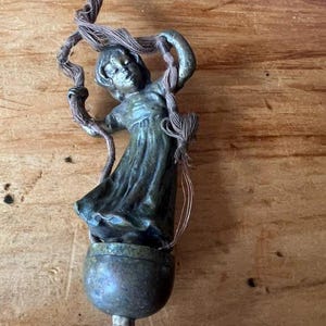 May include: A bronze-toned metal figurine of a person in a flowing dress, positioned above a rounded base. The figure holds a frayed, light brown cord that loops around the arms and extends upward. A small white cylindrical component is visible at the bottom.