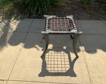 May include: A black metal stool with a woven brown leather seat. The stool has four legs and is sitting on a concrete patio.