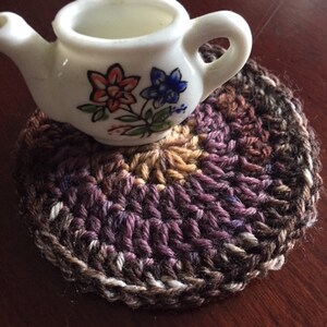 Può includere: Un piccolo teiera bianca con disegni floreali si trova su un sottobicchiere all'uncinetto con filo marrone, viola e bianco.
