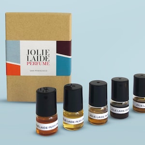 May include: A set of five small brown glass bottles with black caps and white labels that say "Jolie Laide Perfume" in a brown cardboard box. The box has a label that says "Jolie Laide Perfume San Francisco".