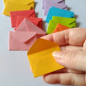 May include: A collection of colourful paper envelopes in various shades of pink, red, orange, yellow, green, blue, and purple. The envelopes are arranged in a semi-circle with a yellow envelope being held in a hand.