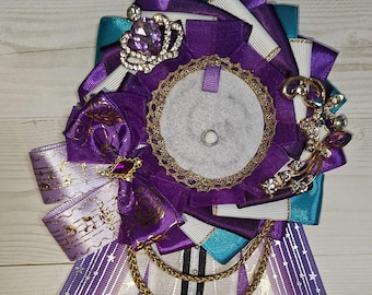 May include: A decorative ribbon rosette in shades of purple, white, and teal, adorned with gold trim, a crown, and a decorative brooch. The design includes a central circle and cascading ribbons, creating a layered effect. A gold chain and jewel accents add to the ornate style.