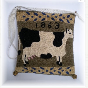 May include: A brown and green pillow with a black and white cow design. The pillow has a white lace trim and two bells on the bottom corners. The year 1863 is embroidered on the pillow.