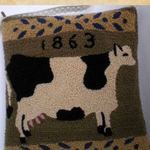 May include: A brown and tan pillow with a black and white cow design. The pillow has a blue and tan border with the year 1863 embroidered on it. The pillow is labeled "Colonial Cow".