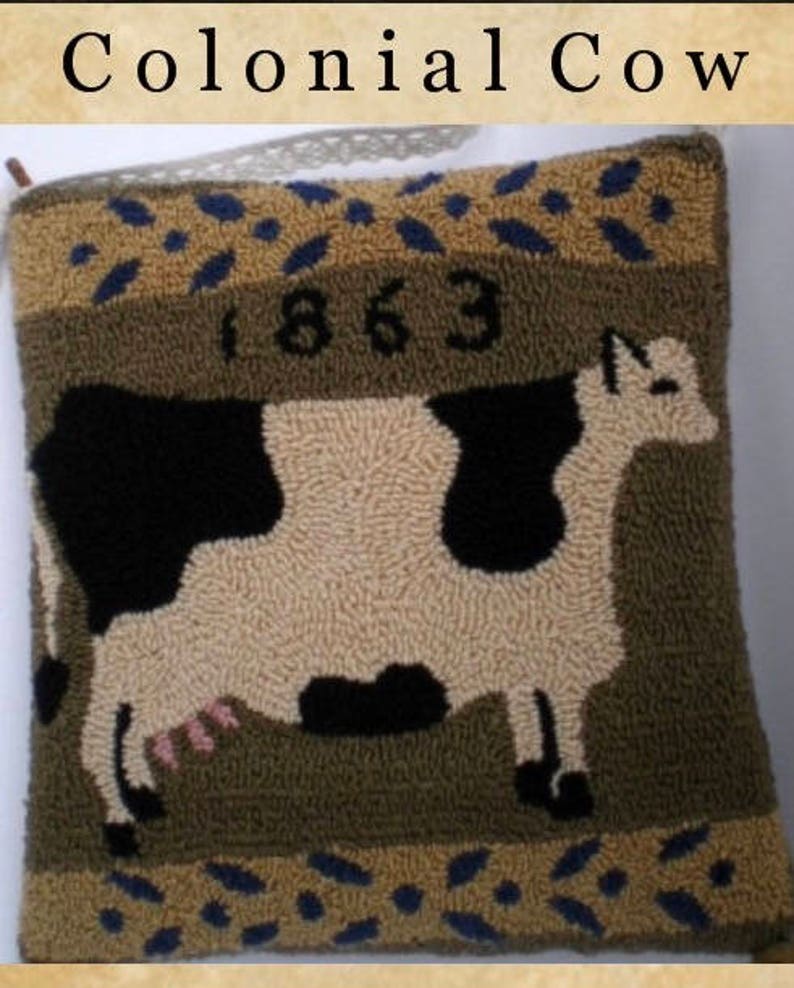 May include: A brown and tan pillow with a black and white cow design. The pillow has a blue and tan border with the year 1863 embroidered on it. The pillow is labeled "Colonial Cow".