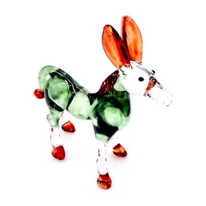 May include: A green glass donkey figurine with orange ears and hooves. The donkey has a white face and a clear glass nose.