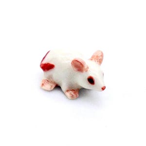 Könnte beinhalten: Kleine Keramik-Mausfigur. Die Maus ist weiß mit rosa Ohren, Füßen und Nase. Sie hat rote Augen und einen roten Fleck auf dem Rücken. Die Figur steht vor einem weißen Hintergrund.