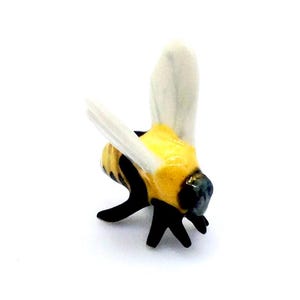 Könnte beinhalten: Eine kleine Keramikfigur einer Hummel mit einem glänzend gelb-schwarz gestreiften Körper, einem dunklen Kopf und zwei aufrechten, durchscheinenden weißen Flügeln. Die Hummel ist in stehender Position dargestellt, wobei schwarze Beine sichtbar sind.