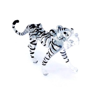 May include: A clear glass tiger figurine with black stripes. The tiger is in a walking pose, with its tail curved upwards. The figurine has a detailed design and is set against a white background.