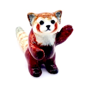 以下が含まれることがあります： 白い胸と顔をした赤いパンダの小さなセラミック製フィギュア。パンダは後ろ足で立ち、前足を上げて手を振っている。