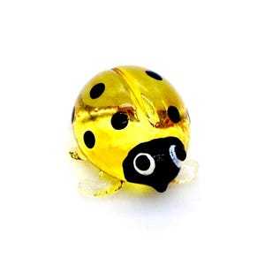 Könnte beinhalten: Eine kleine Marienkäferfigur aus Glas. Der Marienkäfer ist überwiegend gelb mit schwarzen Punkten auf dem Rücken und einem schwarzen Kopf mit weißen Augen. Die Figur hat klare, kleine Flügel und steht vor einem weißen Hintergrund.