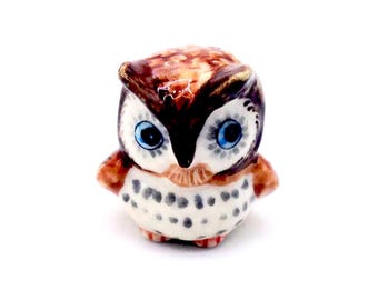Könnte beinhalten: Kleine Eulenfigur aus Keramik mit detailliertem Design. Die Eule hat braune und weiße Federn, große blaue Augen und einen kleinen Schnabel. Die Figur ist ein Dekorationsartikel, wahrscheinlich zur Ausstellung.