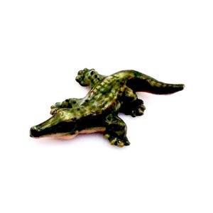 May include: A small, green ceramic alligator figurine with a textured, scaly body. The alligator is lying on its belly with its head turned to the side.