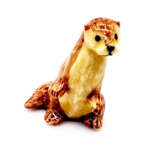 May include: A ceramic otter figurine in shades of brown, tan, and cream. The otter is posed upright, with its front paws resting on its chest. The figurine has a glossy finish and is set against a white background.