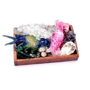 May include: A miniature wooden tray filled with colorful, realistic-looking food items. The tray contains a blue and green crustacean, pink candies, white ice, and small, round, beige objects. The food items are arranged on a white background.