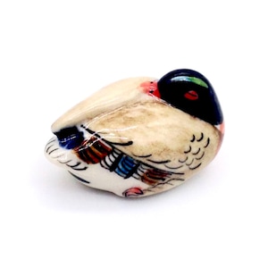 May include: A ceramic duck figurine, resting with its head tucked. The duck has a cream-colored body, a black head with a green sheen, and accents of red, blue, and brown. The figurine is on a white background.