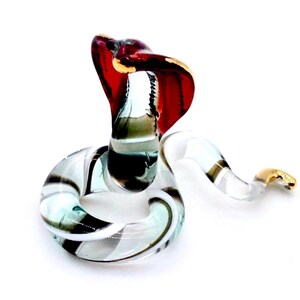May include: A glass cobra figurine with a raised hood. The snake is primarily clear with black, white, and teal accents. The hood is red with gold trim. The snake's body is coiled, and the tail has gold accents.