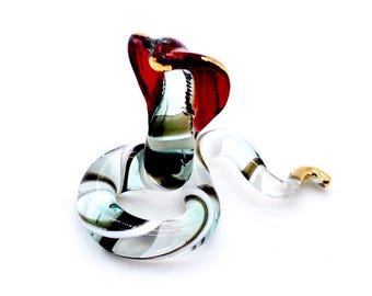 May include: A glass cobra ornament with a raised hood. The snake is mainly clear with black, white, and teal accents. The hood is red with gold trim. The snake's body is coiled, and the tail has gold accents.