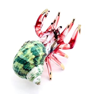 Puede incluir: Una figura de cristal de un cangrejo ermitaño con una concha espiral texturizada en tonos verdes y blancos. El cuerpo está hecho de vidrio rojo translúcido con secciones transparentes y puntas pintadas de dorado en sus diez patas articuladas.