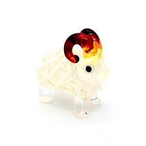 May include: A small glass figurine of a ram with a textured, translucent white body and clear glass legs. The ram features prominent, curved horns that transition from a deep reddish-brown at the base to a bright yellow at the tips, with black eyes.
