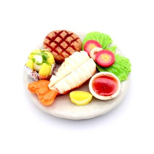 Könnte beinhalten: Ein weißer Teller mit einem gegrillten Steak, einer Ofenkartoffel, einer Zitronenscheibe, einer Beilage mit Garnelen, einer kleinen Schüssel mit roter Sauce und einem Salat aus Salat und Tomatenscheiben.