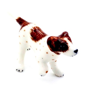May include: A small ceramic figurine of a brown and white spotted dog with brown ears. The dog is standing on all four legs and has its head tilted to the side.