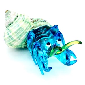 May include: A detailed glass sculpture of a hermit crab. The crab is a vibrant turquoise blue with a green and yellow claw. It emerges from a natural, spiraled seashell with green and white stripes. The background is plain white.
