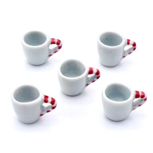 Może przedstawiać: Pięć małych, białych ceramicznych kubków z czerwono-białymi paskami na uchwytach. Miniaturowe kubki ułożone są na białej powierzchni. Kubki te są prawdopodobnie przeznaczone do domku dla lalek lub miniaturowej ekspozycji.