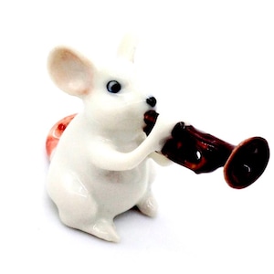 Puede incluir: Una figura de ratón de cerámica blanca tocando una trompeta marrón. El ratón tiene ojos azules y un detalle rojo y rosa en forma de concha en la espalda. La figura está sobre un fondo blanco.
