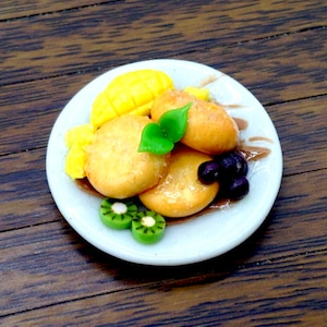 Könnte beinhalten: Ein weißer Teller mit einer gelben Mango, vier kleinen runden frittierten Teigbällchen, zwei grünen Kiwischeiben und ein paar dunkelvioletten Blaubeeren. Der Teller befindet sich auf einer braunen Holzoberfläche.
