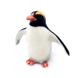 以下が含まれることがあります： 白い体、黒い翼と頭、黄色のトサカを持つ小さなペンギンの置物。 ペンギンは赤いクチバシと足を持ち、翼はわずかに広がっています。 置物は白い背景にあります。