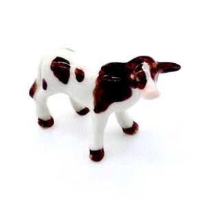 May include: A small ceramic cow figurine. The cow is white with brown spots and patches, and has brown hooves and horns. The figurine is standing on four legs and has a pinkish-white muzzle. The cow is a miniature farm animal.