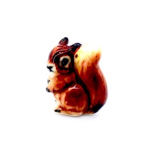 May include: A small, ceramic squirrel figurine. The squirrel is depicted in a sitting position with a fluffy tail. The figurine features a glossy finish with shades of brown, tan, and white. The squirrel's eyes are black.