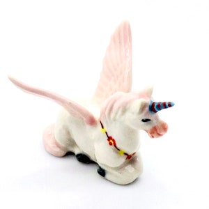 May include: A small white ceramic unicorn figurine with pink wings, a blue and red horn, and a yellow and red flower garland around its neck.