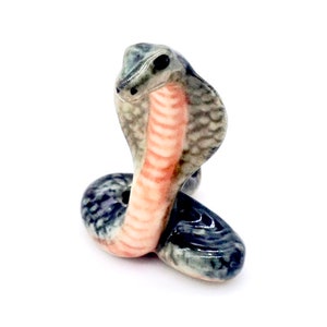 May include: A ceramic cobra figurine in shades of gray, black, and pink. The snake is posed upright, with its hood flared. The figurine has a glossy finish and is set against a white background.