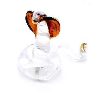 May include: A clear glass cobra figurine with orange and gold accents. The cobra is in a striking pose, with its hood flared and its body coiled. The figurine is set against a white background.