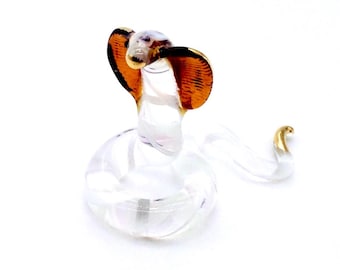 May include: A clear glass cobra figurine with orange and gold accents. The cobra is in a striking pose, with its hood flared and its body coiled. The figurine is set against a white background.