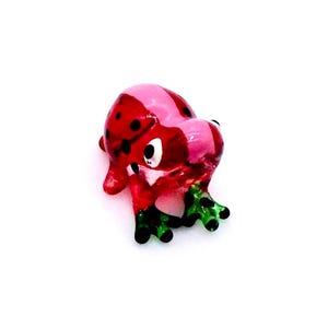 May include: A small, glass ladybug figurine. The ladybug is primarily red with black spots on its back and pink accents. The legs are green, and the eyes are white with black pupils. The figurine is on a white background.