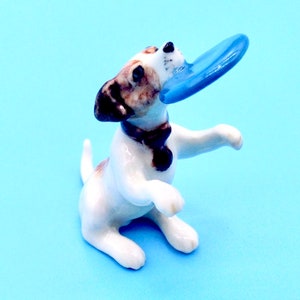 May include: A ceramic figurine of a dog, predominantly white with brown spots, holding a blue frisbee in its mouth. The dog is wearing a dark brown collar and is posed as if playing. The background is a solid light blue.