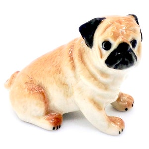 May include: A small ceramic figurine of a pug dog. The pug is sitting with its legs tucked under its body. It has a light brown and white coat with black ears and a black nose.