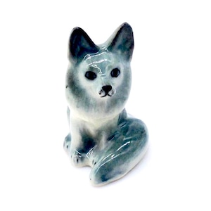 Könnte beinhalten: Eine kleine Keramikfigur eines grauen Fuchses, der mit seinem Schwanz um seinen Körper gewickelt sitzt. Der Fuchs hat schwarze Augen und eine weiße Schnauze.