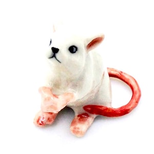 May include: A white ceramic figurine of a small animal, possibly a cat or dog, with a red tail and pink accents on its paws and ears. The figurine has black eyes and a black nose. The animal is sitting upright.